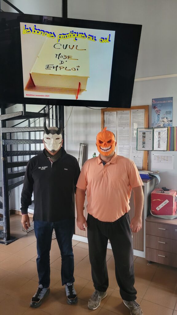 Jean-Paul et Sylvain sont debout sous le grand écran de la maison du planeur. Jean-Paul porte un masque du diable et Sylvain un masque de citrouille